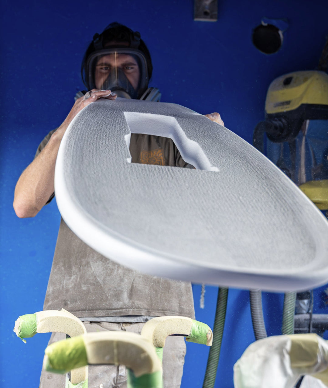 A shaper wearing a full-face mask and protective gear holds a large, white, textured surfboard or deck in a workshop with a bright blue wall background.