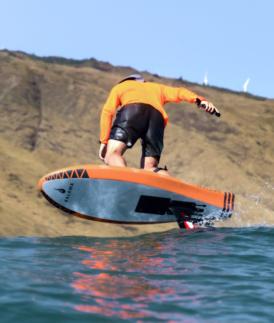 A person in an orange long-sleeve top and black shorts stands on RIIFT-enabled board riding over blue water with a tan hillside and blue sky in the background.