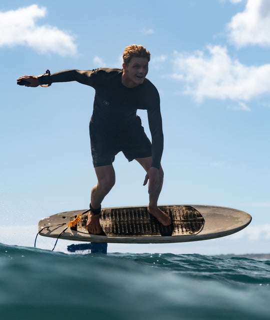 Nathan Van Vuuren in a black wetsuit stands on a RIIFT-enabled foilboard floating on the surface of the ocean under a blue sky with clouds.