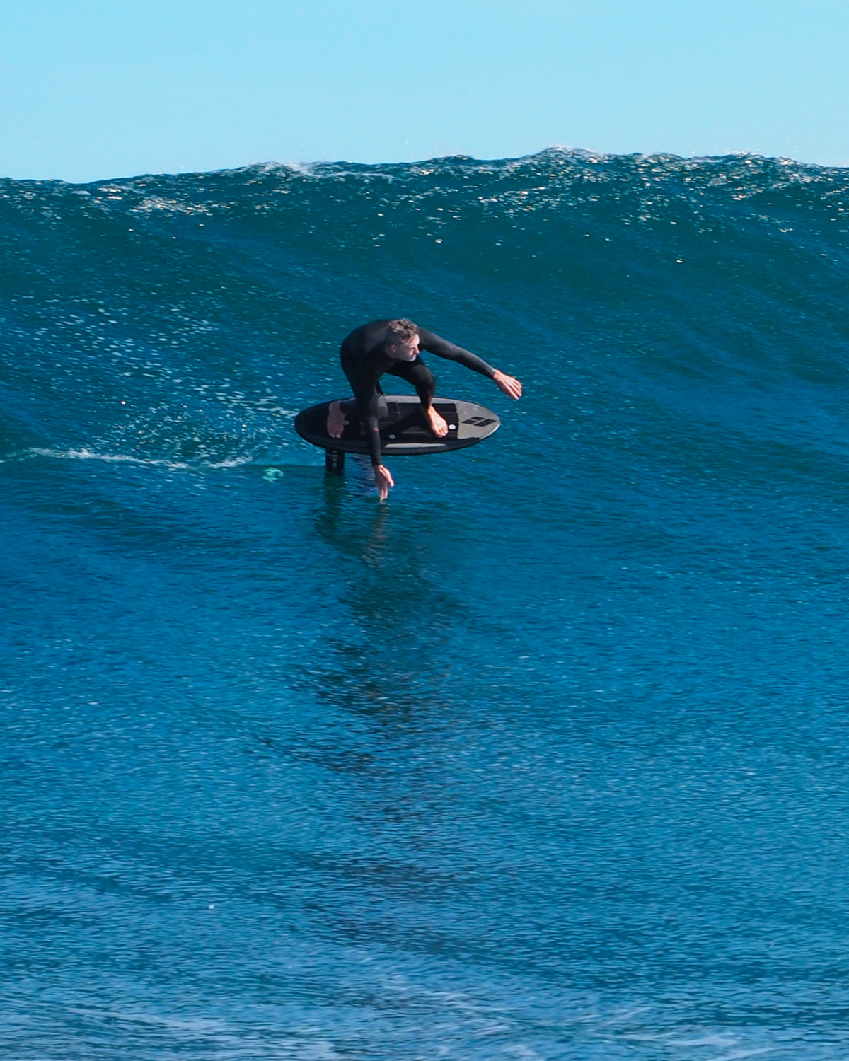 John from RIIFT wears a black wetsuit rides a large blue ocean wave under a pale blue sky.