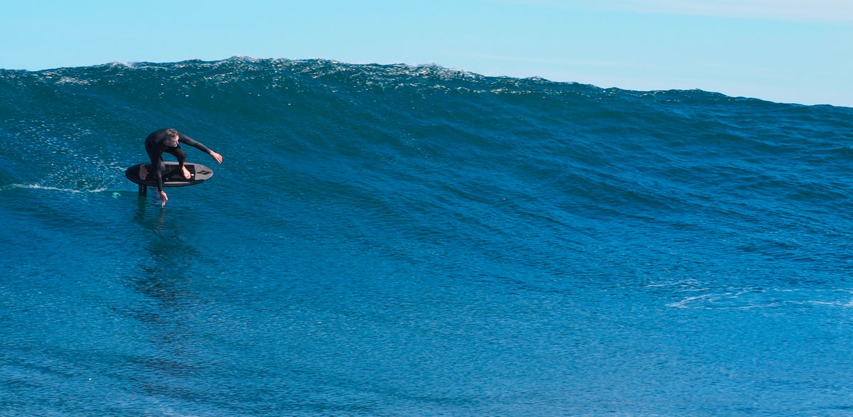 John from RIIFT wears a black wetsuit rides a large blue ocean wave under a pale blue sky.