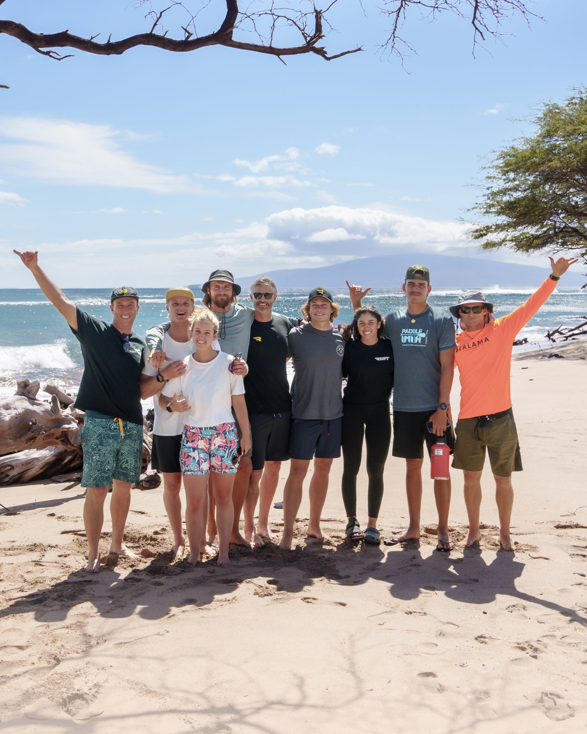 A group of RIIFT friends posing together on a sandy Hawaiian beach with the ocean, mountains, and sky in the background. They are wearing casual beachwear and surf-related apparel, with one person wearing a bright orange long-sleeve shirt and another in a dark green shirt.