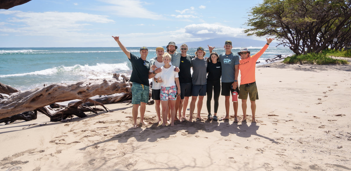 A group of nine RIIFT friends posing together on a sandy beach with the ocean, trees, and sky in the background.