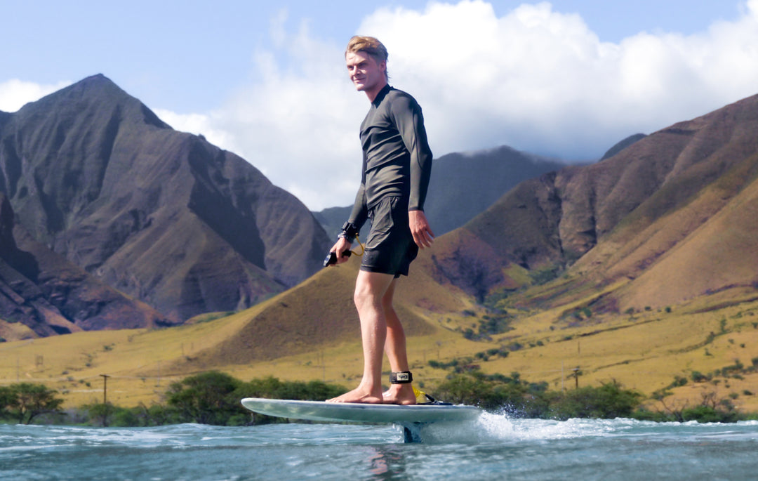 Nathan Van Vuuren stands on a surfboard in a lake with mountains in the background, wearing a black long-sleeve rash guard and black swim trunks.