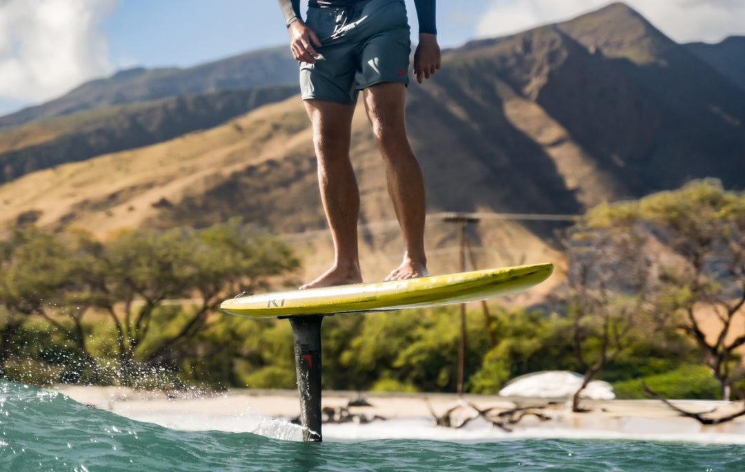 Jarrod Snow wearing a dark teal long-sleeve rash guard and black swim trunks rides a wave on a RIIFT-enabled board in the ocean under a cloudy sky.