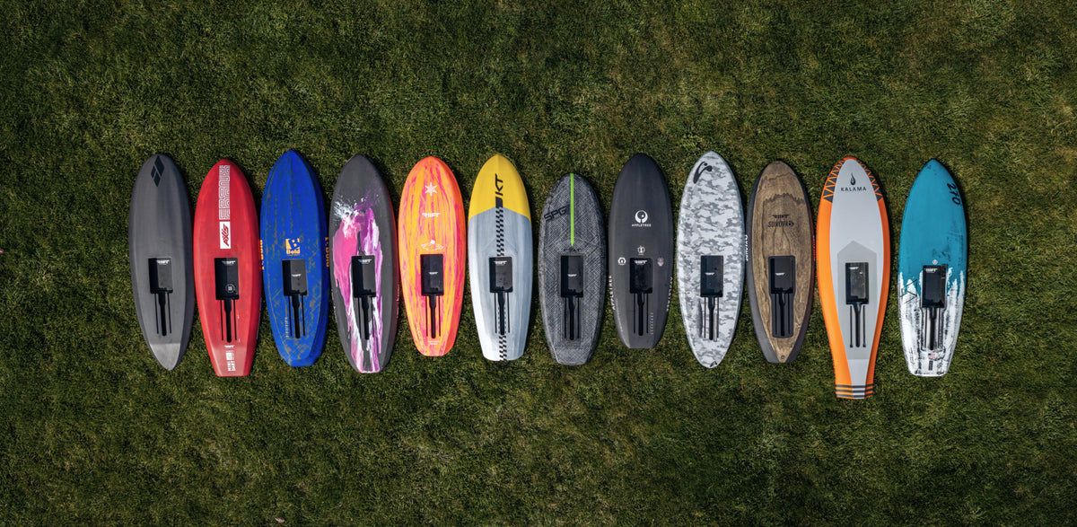 A row of ten RIIFT enabled boards from all the RIIFT shaper partners around the world. They display in various colors and patterns, laid out side-by-side on a green grass lawn.