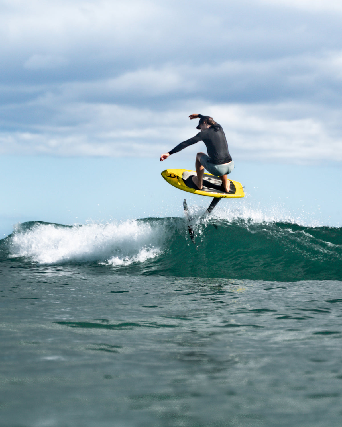 Kane De Wilde n a black wetsuit rides a yellow and black RIIFT-enabled KT board over a breaking ocean wave under a cloudy sky.