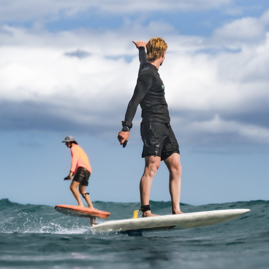 Nathan Van Vuuren stands on a white RIIFT-enabled board in the ocean, with another person in an orange shirt on a board in the background under a cloudy blue sky.