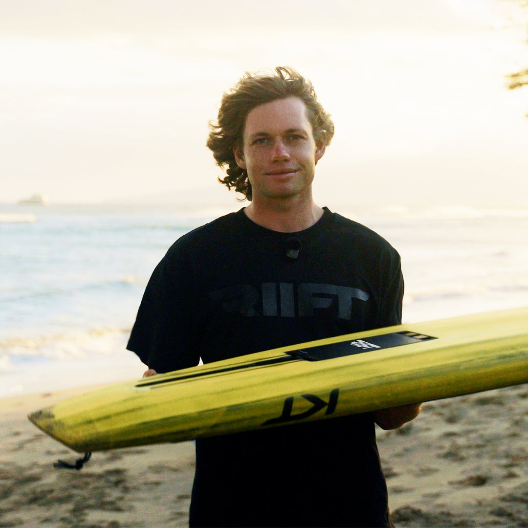 Kane De Wilde standing on a beach holding a yellow KT RIIFT-enabled board. He is wearing a black t-shirt with a dark graphic logo that reads RIIFT.