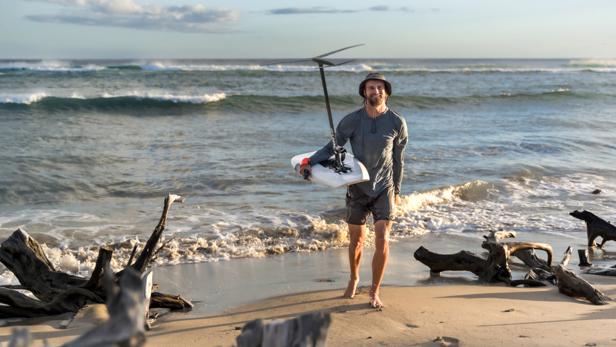 Jarrod Snow on a sandy beach holding a white and black RIIFT-enabled board with a long black pole, with ocean waves and driftwood in the background.