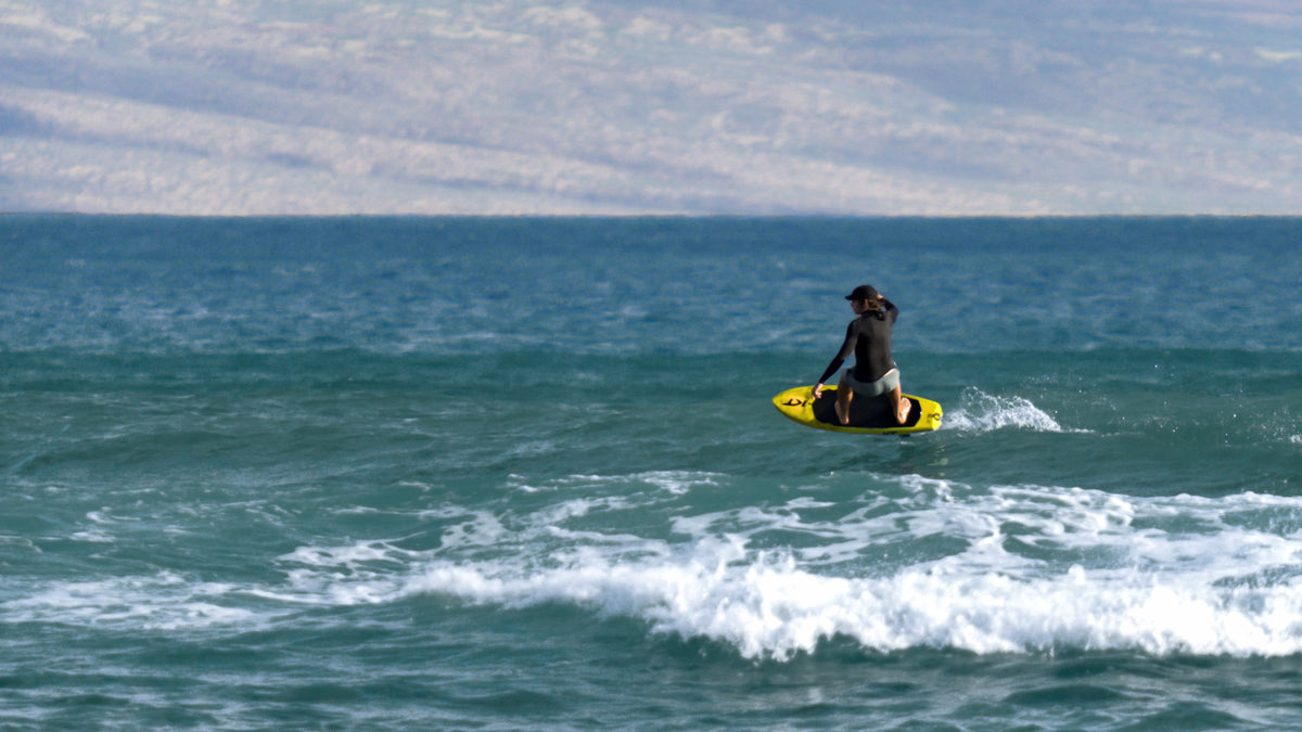 Kane De Wilde in a black wetsuit rides a yellow RIIFT-enabled KT board on the blue ocean with white waves and a hazy mountain range in the background.