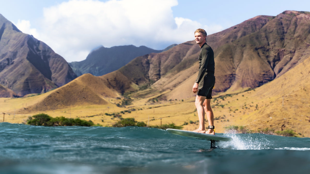 Nathan Van Vuuren in a black wetsuit stands on a RIIFT-enabled board in blue water with a dramatic mountain landscape in the background in Hawaii.