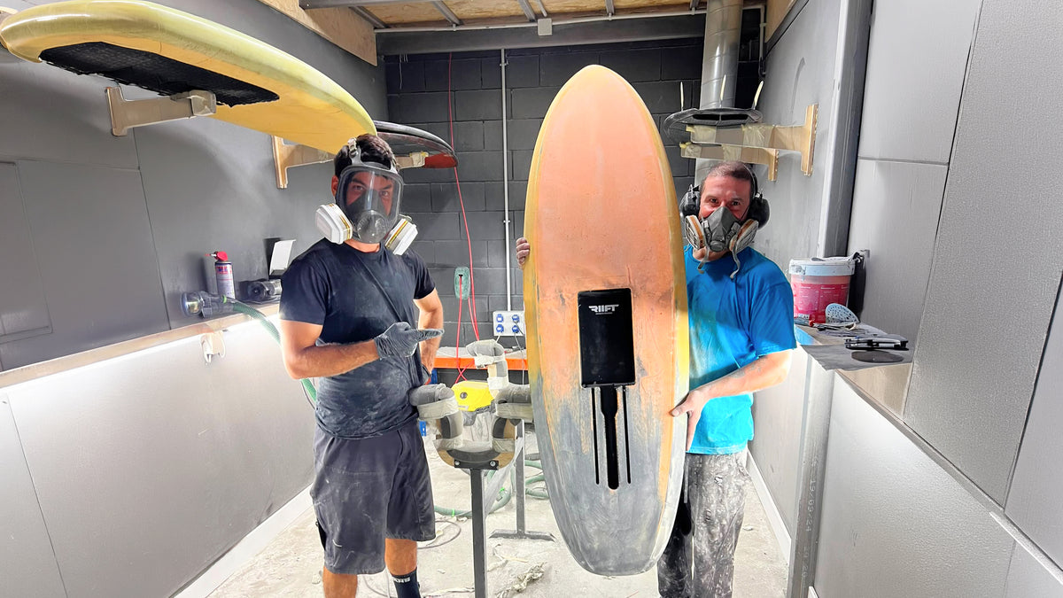 Two men wearing respirator masks and protective clothing stand in a board shaping workshop holding a large surfboard with RIIFT tracks. Another surfboard hangs on the wall in the background.